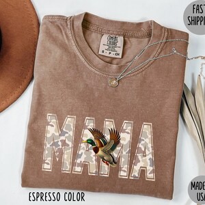 May include: Espresso-colored t-shirt with the word "MAMA" in a camouflage pattern, featuring a detailed illustration of a duck. The shirt has a crew neck and short sleeves. A silver necklace with a pendant is draped on the shirt.