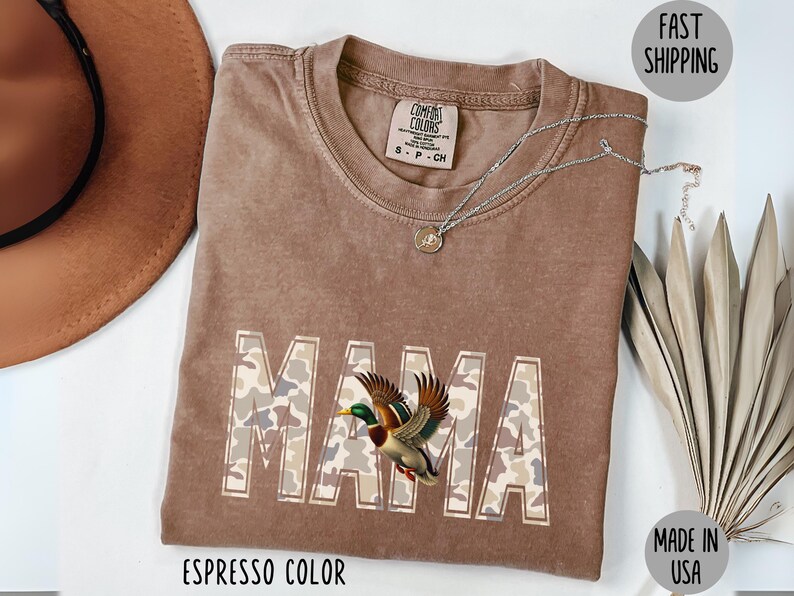 May include: Espresso-colored t-shirt with the word "MAMA" in a camouflage pattern, featuring a detailed illustration of a duck. The shirt has a crew neck and short sleeves. A silver necklace with a pendant is draped on the shirt.
