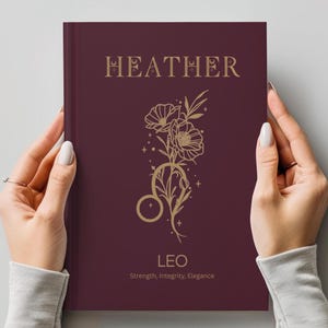 May include: A dark maroon hardcover book featuring the name HEATHER in gold lettering at the top. Below, a gold line art illustration of flowers and the Leo zodiac symbol is centered. The word LEO and the text Strength, Integrity, Elegance appear below.