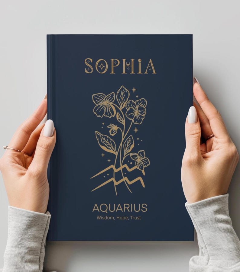 May include: A dark blue hardcover journal featuring gold foil lettering and line art. The cover displays the name SOPHIA at the top, a floral illustration with the Aquarius zodiac symbol below it, and the text AQUARIUS Wisdom, Hope, Trust.