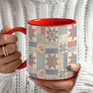 May include: A white ceramic mug with a red interior and handle. The exterior features a patchwork quilt design with blue, red, and floral patterns. The mug is held by a person wearing a white sweater.
