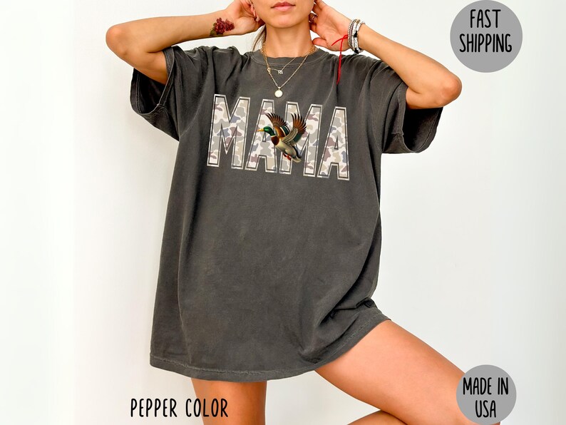 May include: Dark grey t-shirt with the word "MAMA" in camouflage print, featuring a duck graphic. The shirt is a pepper color and made in the USA.