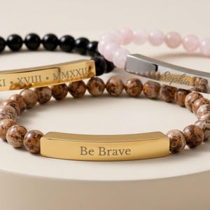 May include: Three beaded bracelets featuring metal bars. One bracelet has brown speckled beads and a gold bar engraved with "Be Brave". Another has black beads and a gold bar with "XVIII • MMXXXII". The third has light pink beads and a silver bar with "Sophia".