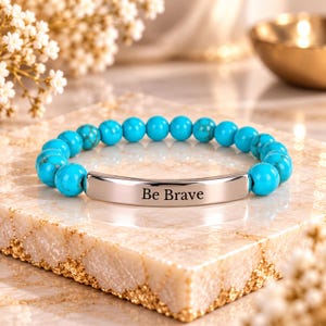 May include: A stretch bracelet featuring round, bright turquoise beads with dark veining and a central, polished silver-tone metal bar engraved with the text Be Brave in a clean, black, sans-serif font.