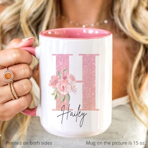 May include: White ceramic mug with a pink interior and handle. The mug features a large, glittery pink letter "H" with a floral bouquet and the name "Hailey" in script. The mug is 15 oz.