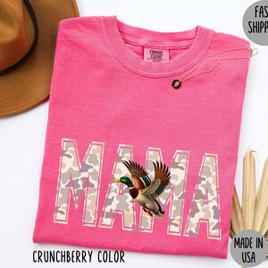 May include: A pink t-shirt with the word "MAMA" in a camouflage pattern, featuring a flying duck graphic. The shirt is labeled "Crunchberry Color" and "Made in USA". A gold necklace and a brown hat are also visible.