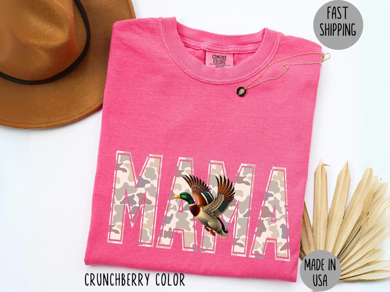May include: A pink t-shirt with the word "MAMA" in a camouflage pattern, featuring a flying duck graphic. The shirt is labeled "Crunchberry Color" and "Made in USA". A gold necklace and a brown hat are also visible.