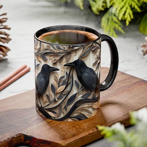 May include: A black coffee mug with a detailed relief design featuring two black crows perched among stylized leaves and branches. The mug has a black handle and rim, and is filled with hot coffee. The mug is placed on a wooden cutting board.