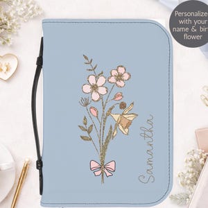 Può includere: Una copertina di libro azzurra con un design floreale. La copertina presenta un bouquet ricamato di fiori rosa, un narciso e il nome "Samantha" in corsivo. Un manico nero è attaccato al lato. Il testo sull'immagine dice "Personalizzato con il tuo nome e fiore di nascita."