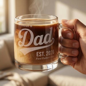Puede incluir: Una taza de café de vidrio transparente llena de una bebida marrón oscuro y crema. La taza tiene las palabras "Promoted to Dad" y "EST. 2026 First Father's Day" grabadas en la parte delantera. Sale vapor de la taza.
