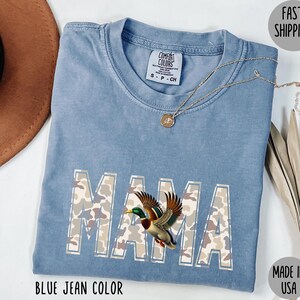 May include: Blue jean colored Comfort Colors t-shirt with the word "MAMA" in camouflage print and a duck graphic. A gold necklace rests on the shirt. The shirt is made in the USA.