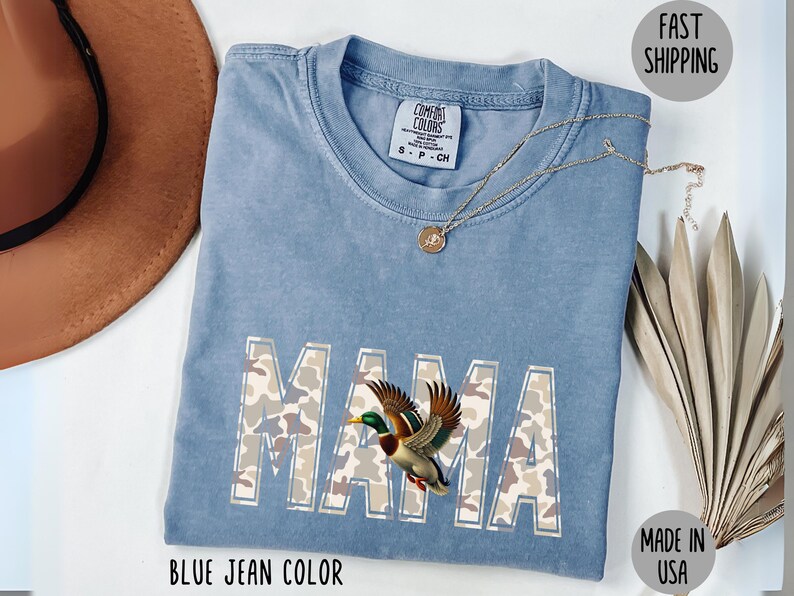 May include: Blue jean colored Comfort Colors t-shirt with the word "MAMA" in camouflage print and a duck graphic. A gold necklace rests on the shirt. The shirt is made in the USA.