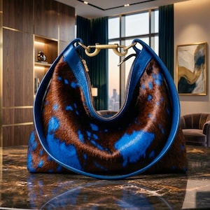 May include: A crescent-shaped handbag featuring a brown and blue cowhide pattern with a smooth blue leather trim. The bag includes a gold-toned metal hardware clasp at the top center, connecting the two sides of the circular frame.
