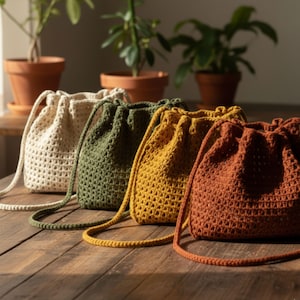 Puede incluir: Cuatro bolsas de crochet con cordón hechas a mano en crema, verde oliva, amarillo mostaza y naranja óxido. Cada bolsa tiene un cordón a juego y un diseño de tejido abierto texturizado. Las bolsas se exhiben sobre una superficie de madera.