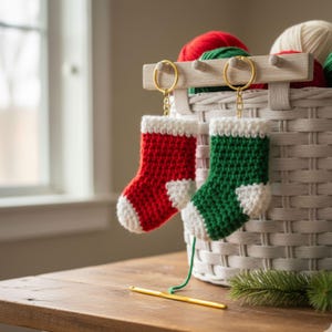 May include: Two miniature crochet Christmas stockings, one red and one green, with white accents, hang from gold keyrings. A basket filled with yarn and a crochet hook are also visible, suggesting a handmade theme.
