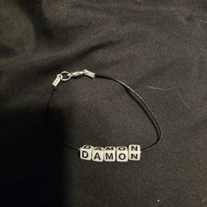 May include: A black cord bracelet with white square beads spelling out the name "DAMON". The bracelet has a silver clasp.