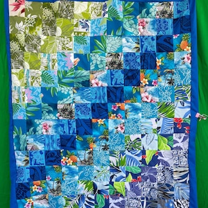 May include: A patchwork quilt with a blue border, featuring a variety of blue and green floral and tropical print squares. The quilt showcases a range of patterns, including hibiscus flowers, palm trees, and tropical leaves. The quilt is hanging against a green background.