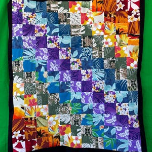 May include: A colorful patchwork quilt with a black border, featuring squares of fabric in various floral and tropical patterns. The quilt showcases a vibrant mix of colors, including purple, blue, yellow, and red, creating a visually appealing design. The quilt is hanging against a green background.