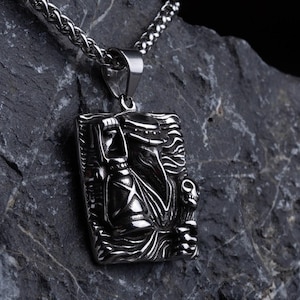 May include: A silver pendant necklace with a rectangular pendant displaying a detailed relief design. The pendant is suspended from a chain and is placed on a dark grey rock against a black backdrop. The design seems to be a stylized figure.