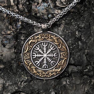 Könnte beinhalten: Eine silberne Halskette mit Anhänger, mit einem kreisförmigen Anhänger. Der Anhänger hat einen goldfarbenen Rand und ein detailliertes, symmetrisches Design. Die Halskette hängt an einer silbernen Kette vor einem dunklen, strukturierten Hintergrund.