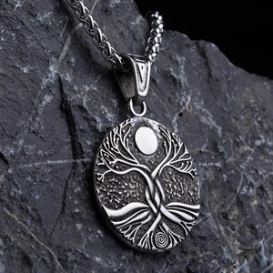 Puede incluir: Collar con colgante plateado con un diseño de árbol de la vida. El colgante ovalado muestra un árbol detallado con un sol o una luna en la parte superior. El collar se exhibe sobre una roca gris oscura y texturizada sobre un fondo negro.