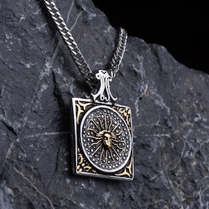 以下が含まれることがあります： 太陽のデザインが施された、正方形のペンダントが付いたシルバーのネックレス。太陽は、精巧なディテールが施されたシルバーのフレームで囲まれています。ネックレスは、黒い背景に対して、濃い灰色の岩の上に展示されています。