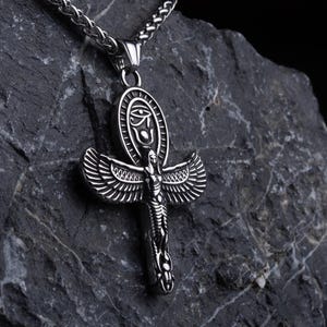 May include: A silver Ankh pendant necklace with intricate detailing, displayed on a dark grey rock against a black backdrop. The Ankh features an eye symbol and winged elements.