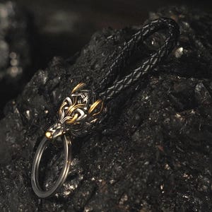May include: A keychain featuring a detailed metal wolf head pendant with gold and silver accents. The pendant is attached to a braided black leather strap and a silver key ring. The background is a dark, textured surface.
