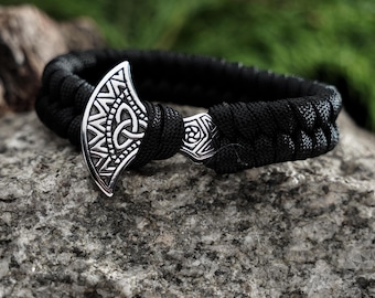 May include: Black braided bracelet with a silver axe-shaped clasp. The clasp features intricate Celtic knot and geometric designs. The bracelet is displayed on a gray stone surface with a blurred green background, suggesting an outdoor setting. The bracelet is a piece of jewelry.