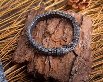 May include: A silver bracelet with a snake design. The bracelet is made of metal and has a textured surface. The bracelet is lying on a piece of wood.