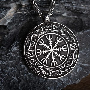 May include: A silver-colored pendant necklace featuring a circular pendant with a detailed Viking symbol design. The pendant is suspended from a chain and rests on a dark, textured stone surface. The pendant's design is a prominent, central focus.