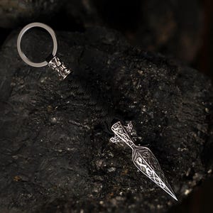 May include: A silver-coloured keychain with a decorative spearhead design. The spearhead is intricately detailed with patterns and attached to a black woven cord. The keychain includes a silver ring for attaching keys.