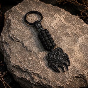 Puede incluir: Un llavero negro con un mango de paracord trenzado y un colgante de metal en forma de pata de oso con detalles intrincados de nudos celtas. El colgante está unido a una anilla metálica circular para sujetar las llaves.