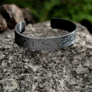 Puede incluir: Un brazalete plateado con un intrincado diseño de nudos celtas. El brazalete abierto descansa sobre una piedra gris texturizada, con un fondo verde borroso. El brazalete presenta patrones detallados y un acabado oscuro oxidado.
