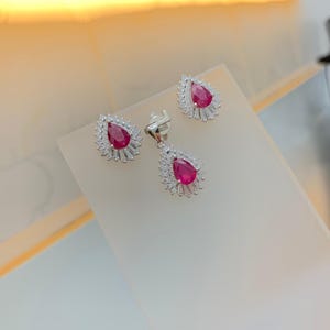 Könnte beinhalten: Ein Schmuckset mit Anhänger und passenden Ohrringen. Jedes Stück hat einen tropfenförmigen, tiefroten Edelstein, der von einem Kranz aus kleineren, klaren Steinen umgeben ist. Das Set wird auf einer weißen Oberfläche präsentiert.