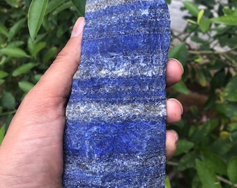 May include: A polished lapis lazuli crystal point. The stone is a deep blue color with streaks of gray and white. The crystal is held in a hand, with green foliage in the background. The crystal is approximately 5 inches tall.