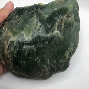 Puede incluir: Una piedra verde grande y tosca con una superficie texturizada. La piedra presenta diferentes tonos de verde, desde el verde oscuro del bosque hasta áreas más claras, casi translúcidas. La forma general es irregular, con un aspecto natural y sin pulir.