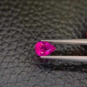 Puede incluir: Una vibrante gema rosa intenso en forma de pera es sostenida por unas pinzas plateadas. Las facetas de la gema reflejan la luz, mostrando su brillo. El fondo es una superficie oscura y texturizada.