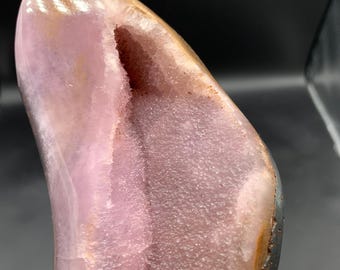 May include: A polished, natural crystal geode with a cavity. The geode features a gradient of colors, including shades of pink, brown, and white. The interior of the geode is filled with small, sparkling crystals.