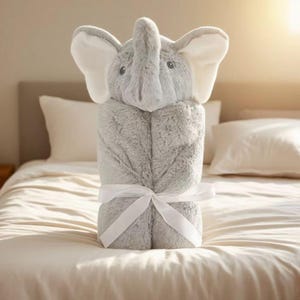 May include: A gray plush elephant hooded baby blanket tied with a white ribbon. The blanket has white inner ears and a detailed elephant face. The blanket is folded and displayed on a bed with white pillows and a cream-colored comforter.