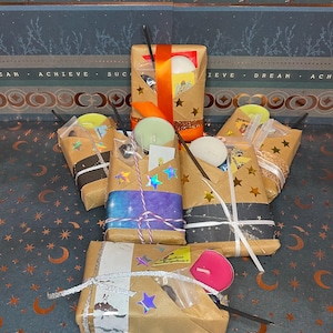 May include: Several wrapped packages with candles, incense, and decorative stars. The packages are wrapped in brown paper and tied with ribbons and twine. The background has text that reads "IN THE MOONLIGHT THERE IS MAGIC."