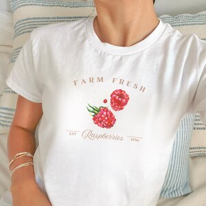May include: White t-shirt with a raspberry design. The shirt features the text "FARM FRESH" above two red raspberries with green leaves. Below the raspberries is the word "Raspberries" and the year "1956".