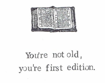 May include: A black and white illustration of an open book with the text "You're not old, you're first edition."