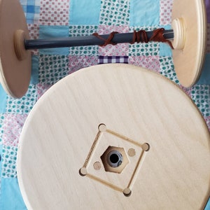 Puede incluir: Un ovillador de hilo de madera con dos extremos de madera redondos y grandes conectados por una varilla gris oscura. El hilo marrón está enrollado alrededor de la varilla. El ovillador está sobre un fondo de colcha de retazos.