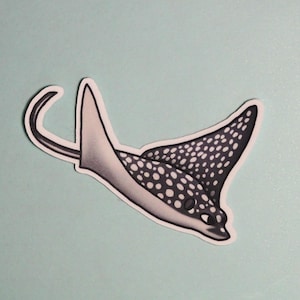May include: A sticker featuring a stylized illustration of an eagle ray. The ray is depicted in shades of gray and white, with a pattern of white spots on its wings. The sticker has a black outline and is set against a light blue background.