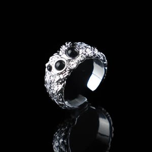 Könnte beinhalten: Ein silberfarbener Ring mit strukturierter Oberfläche und drei schwarzen Edelsteinen. Der Ring ist unten offen und spiegelt sich auf der schwarzen Oberfläche darunter.