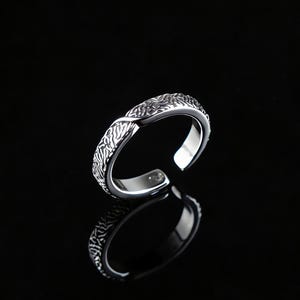 Puede incluir: Un anillo ajustable plateado con un patrón texturizado similar a un tejido. El diseño abierto permite ajustar la talla. El anillo se muestra sobre un fondo negro, con su reflejo visible debajo.
