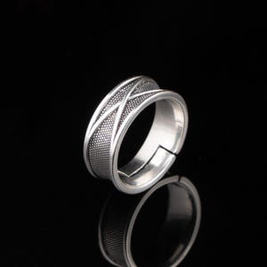 Op de afbeelding: Een zilveren ring met een getextureerde band en een open verstelbaar ontwerp. De band heeft een gestippeld patroon en een kruisontwerp. De ring wordt gepresenteerd op een reflecterend zwart oppervlak.