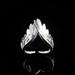 Op de afbeelding: Een zilverkleurige ring met een vleugelontwerp. De ring heeft twee gestileerde vleugels die elkaar aan de bovenkant ontmoeten en een V-vorm creëren. De ring staat tegen een zwarte achtergrond, met een reflectie eronder zichtbaar.