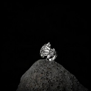 Peut inclure: Une bague en argent avec un design orn&eacute; inspir&eacute; de la nature, avec des feuilles et des lignes fluides. La bague est pos&eacute;e sur un fond sombre, soulignant ses d&eacute;tails complexes et son savoir-faire. La bague semble &ecirc;tre en argent sterling.